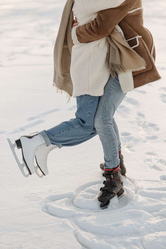 Es sind zwei Menschen bis zu den Schultern zu sehen, welche sich in winterlichen Klamotten hochspringend umarmen. Sie stehen auf Schnee und tragen Schlittschuhe. Ihr Outfit und Aktivität passt zu den First Date Looks und Ideen.