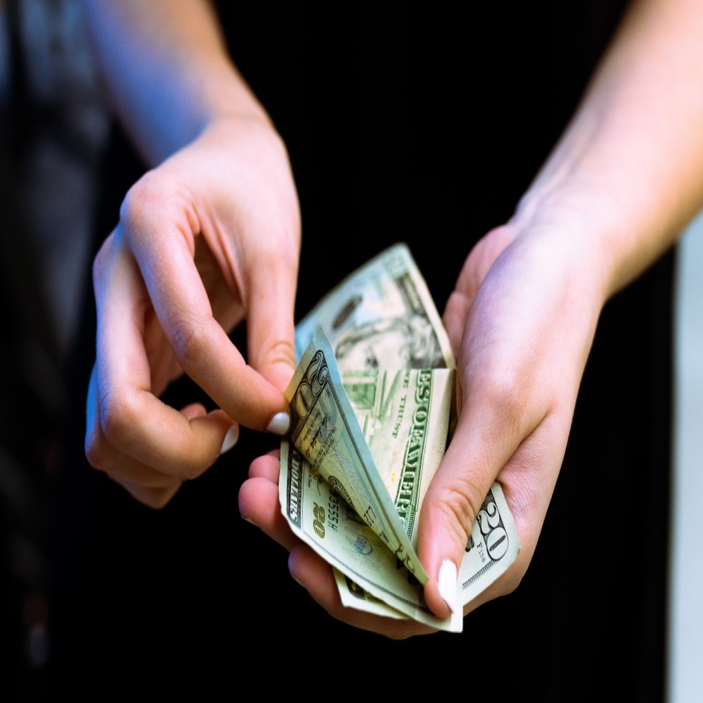 Zwei weibliche Hände mit kurzen, weiß lackierten Fingernägeln halten ein paar Dollarscheine in die Kamera und blättern diese durch. Es kann Geld durch den ARDELL Brand Spot gewonnen werdne.