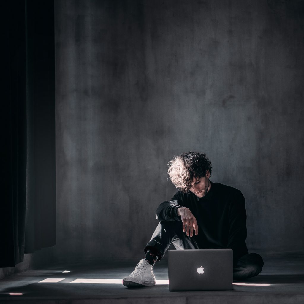 Ein junger Mann sitzt in einem dunklen, schlichten Raum vor seinem Macbook. Er ist schwarz angezogen und schickt online seine Bewerbung ab. 