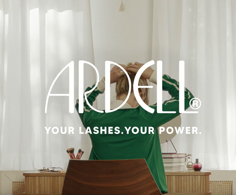 Der ARDELL Slogen "Your Lashes.Your Power." ist im Vordergrund. Im Hintergrund befindet sich eine Frau am Schreibtisch, welche sich die Haare zusammen bindet. Dies ist auch im Werbespot zu sehen.