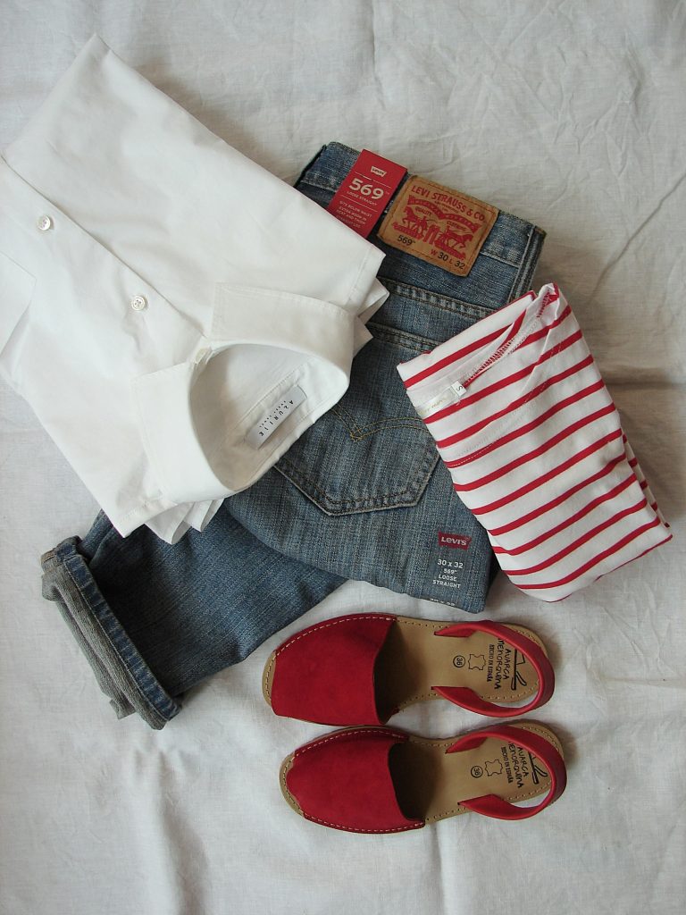 Auf einem Leinentuch liegen eine Bluse, ein paar Jeans und ein rot-weiß gestreiftes Langarmshirt. Daneben stehen rote Sandalen.